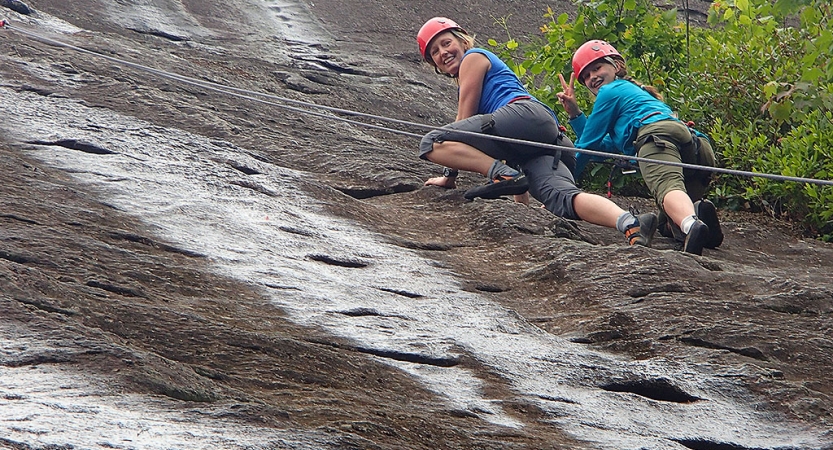 A parent and child look down and smile at the camera while climbing a rock wall. They are both wearing safety gear and are secured by ropes.